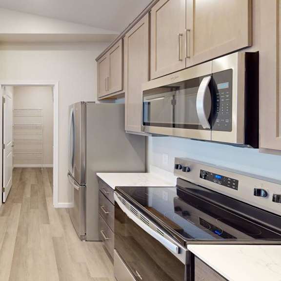 a modern galley-style kitchen here at autumn haus featuring smooth white countertops, warm-toned wood cabinetry with sleek hardware, a stainless steel sink, dishwasher, refrigerator, oven, and built-in microwave, wood-style flooring, and a bright, open layout with a pantry visible at the end of the kitchen for added storage and functionality.
