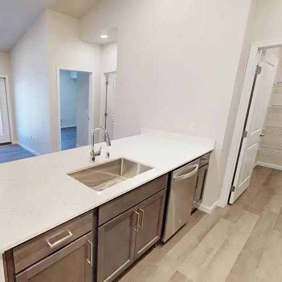 a bright open kitchen here at autumn haus featuring a long white island with an undermount sink and modern faucet, warm wood cabinetry with sleek hardware, a built-in dishwasher, wood-style flooring, recessed ceiling lighting, and an open view into the living area, with a nearby pantry offering ample storage and a clean, airy layout.