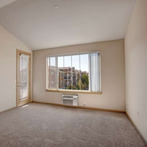 a spacious, empty living room here at the vintage with soft beige carpet, light neutral walls, and a large window bringing in natural light. a glass door with blinds leads outside, and a wall-mounted air conditioner sits below the window.