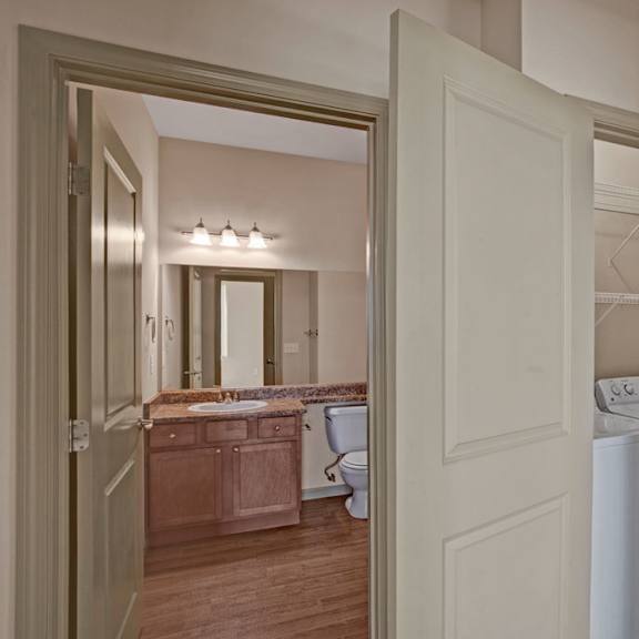 a convenient laundry area here at the vintage with a full-size washer and dryer tucked into a closet with built-in shelving. a nearby bathroom features a wooden vanity with a speckled countertop, large mirror, and bright overhead lighting.