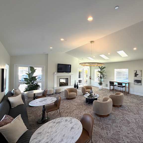 a spacious resident lounge here at the vintage with patterned carpet, large windows bringing in natural light, and multiple seating areas with round tables and upholstered chairs. a cozy fireplace with a mounted tv and modern lighting create a welcoming gathering space.