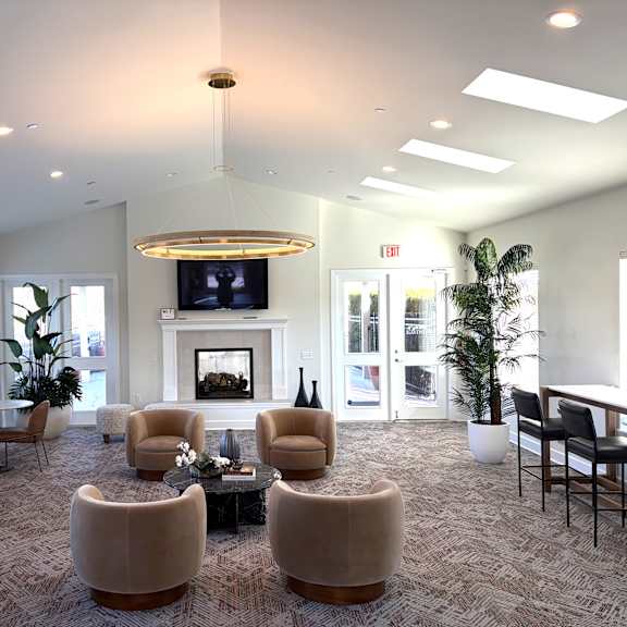 a bright resident lounge here at the vintage with patterned carpet, cozy seating around a round coffee table, and a fireplace with a mounted tv. large windows, tall plants, and modern lighting create a warm and welcoming gathering space.
