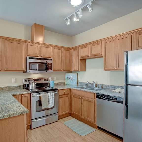 a spacious kitchen here at the vintage with light wood cabinets, speckled granite-style countertops, and stainless steel appliances including a refrigerator, microwave, stove, and dishwasher. a double sink and bright overhead lighting create a clean, modern feel.