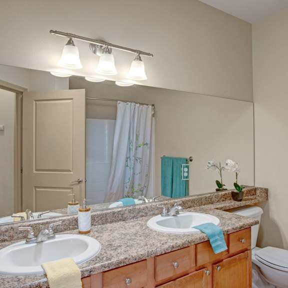 a clean bathroom here at the vintage with a wide mirror, double sinks, and a speckled granite-style countertop. light wood cabinets add warmth, while bright vanity lighting and a shower with a white curtain create a fresh, inviting space.