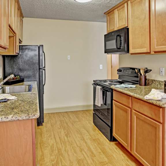 a bright kitchen here at whispering cedars with light brown cabinets, granite countertops, and black appliances, featuring a stainless steel sink, built-in microwave, and wood-style flooring, creating a clean, modern space with a warm, welcoming feel.