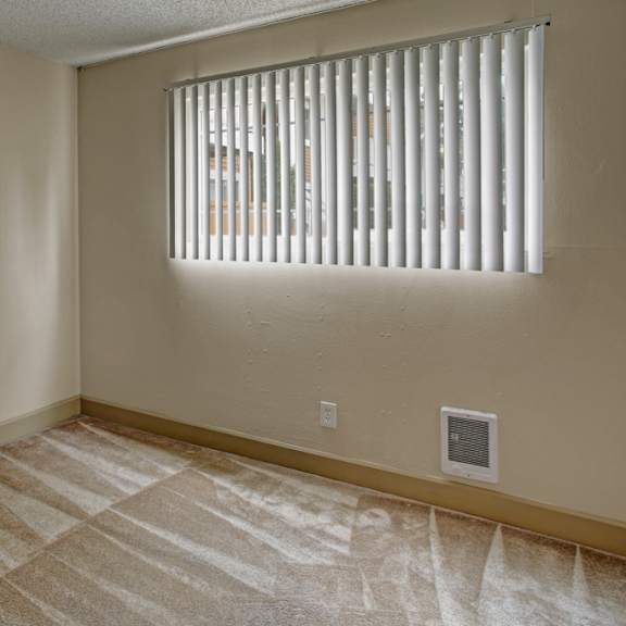 a cozy, empty bedroom here at whispering cedars with soft carpet, neutral walls, and a window with vertical blinds, allowing natural light to fill the space and create a bright, comfortable atmosphere.