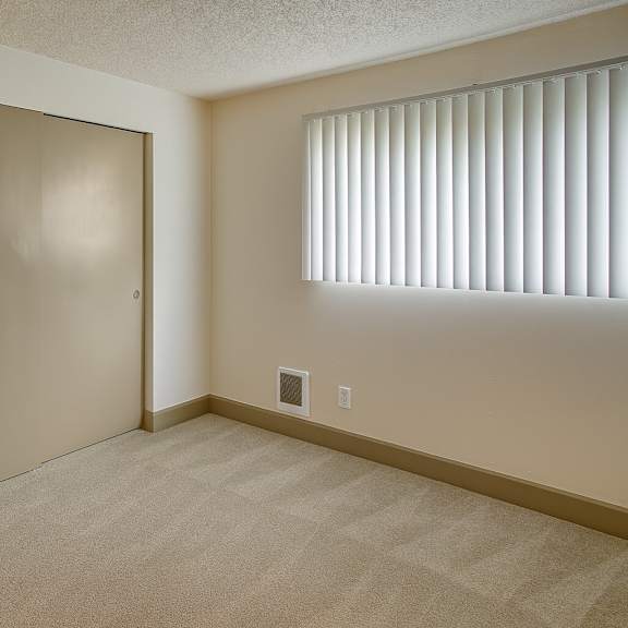a bright, empty bedroom here at whispering cedars with soft carpet, neutral walls, and a window with vertical blinds, featuring a spacious sliding door closet and a clean, comfortable layout.
