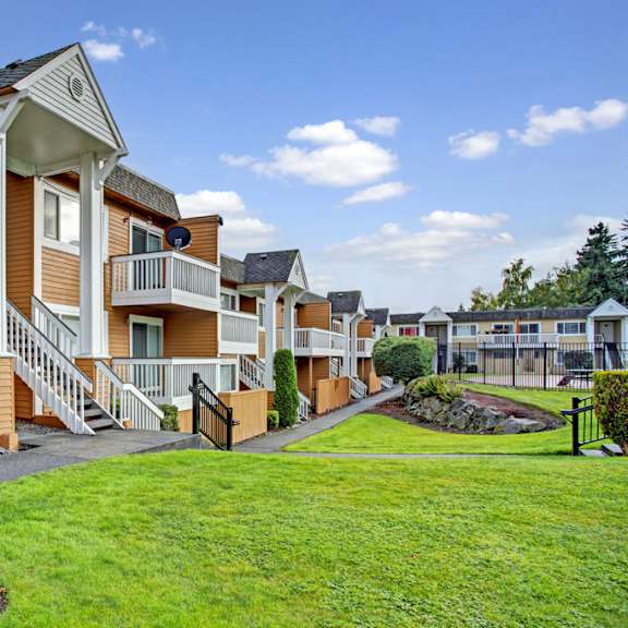 a charming exterior here at whispering cedars with well-kept green lawns, landscaped shrubs, and inviting apartment buildings featuring private balconies, exterior staircases, and a peaceful community setting under a bright open sky.