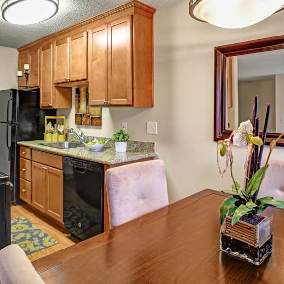a cozy kitchen here at whispering cedars with light brown cabinets, granite countertops, and black appliances, featuring a stainless steel sink, dishwasher, and adjacent dining area with a wooden table and decorative mirror, creating a warm, inviting feel.