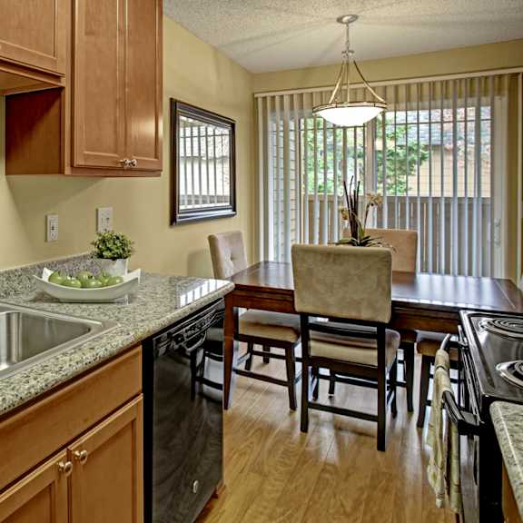 a bright kitchen here at whispering cedars with light brown cabinets, granite countertops, and a stainless steel sink, featuring black appliances and an adjacent dining area with a wooden table, cushioned chairs, and sliding glass doors bringing in natural light.