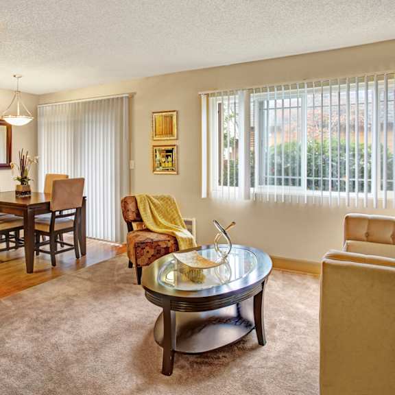 a cozy living room here at whispering cedars with beige carpet, large windows with vertical blinds, and soft natural light, featuring comfortable seating and a glass coffee table, with an adjacent dining area and sliding glass door leading outside.