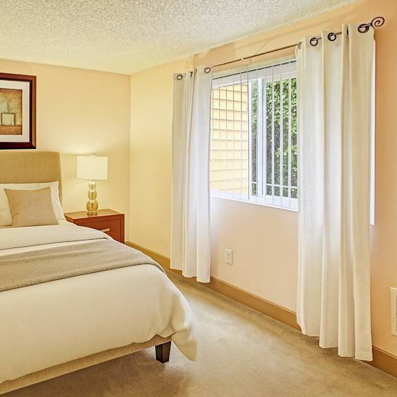 a bright bedroom here at whispering cedars with beige carpet, cream walls, and large windows with white curtains, featuring a comfortable bed with neutral bedding, matching bedside tables with lamps, and a warm, inviting atmosphere.