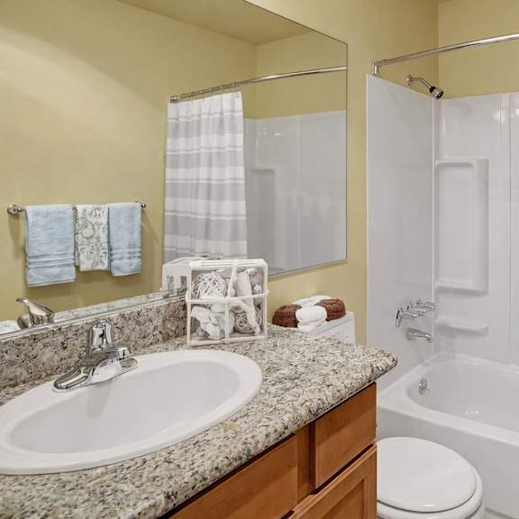a clean bathroom here at whispering cedars with a bathtub-shower combo, white surround, and striped shower curtain, featuring a granite countertop vanity with a large mirror, white sink, and warm wood cabinets, creating a bright, polished feel.