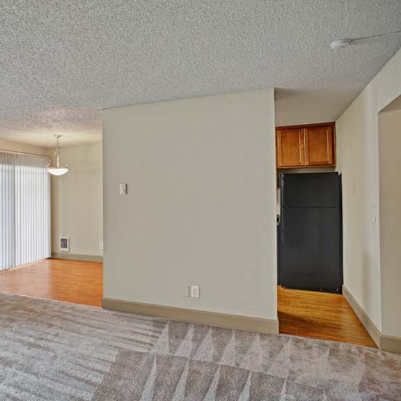 a spacious, empty living room here at whispering cedars with soft carpet, large windows with vertical blinds, and abundant natural light, opening to a dining area with wood-style flooring and a sliding glass door, creating a bright, open layout.