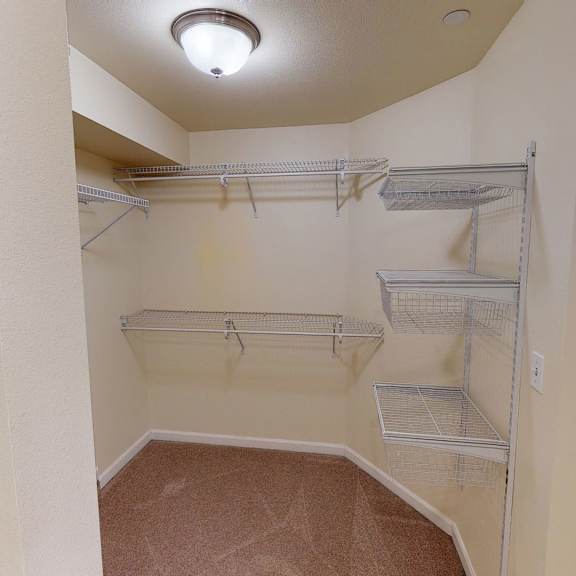 a spacious walk-in closet with beige walls and carpeted floor, featuring metal wire shelving and baskets. soft, warm lighting from a ceiling fixture. located at admirals cove apartments.
