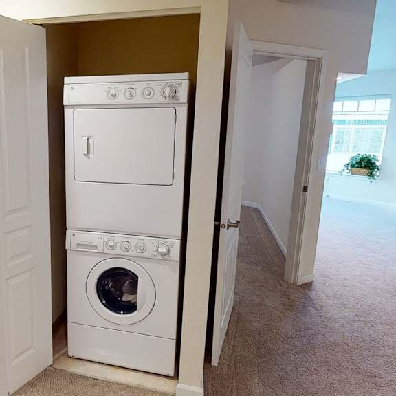 stacked washer and dryer in a small open closet, with adjacent carpeted hallway and bright room with a large window and potted plant in the background. located at admirals cove apartments.