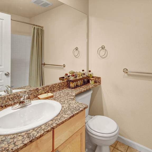 compact bathroom with beige walls, white sink, and toilet. granite countertop holds decorative items. neutral shower curtain and metal towel bars. clean and tidy ambiance. located at admirals cove apartments.