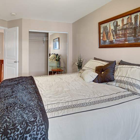cozy admirals cove apartment bedroom with a neatly made bed featuring patterned sheets and pillows. a large painting hangs above the bed, and a closet with mirrored doors is open. soft neutral tones create a calm ambiance.