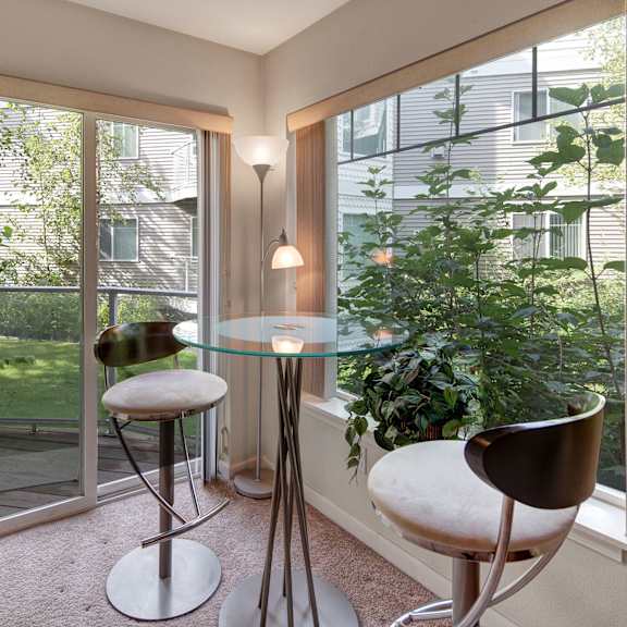 modern dining nook with a glass table and two sleek metal stools, beside large windows overlooking lush greenery. bright, serene atmosphere. located at admirals cove apartments.