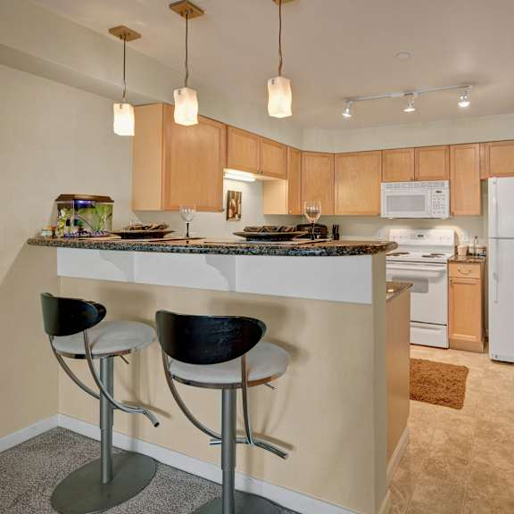 compact kitchen with light wood cabinets, white appliances, and a small dining bar with two stools. hanging lights illuminate the space, and a fishbowl sits on the counter at admirals cove apartments