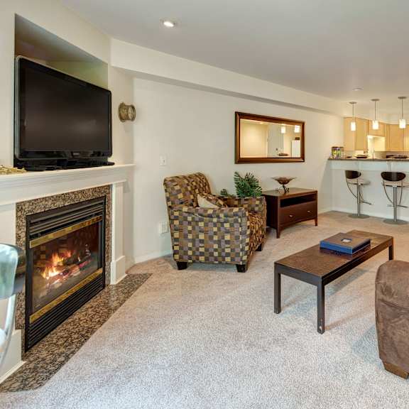 cozy living room at admirals cove with a lit fireplace, wall-mounted tv, and patterned armchair. open kitchen with bar stools and modern appliances. warm tones.