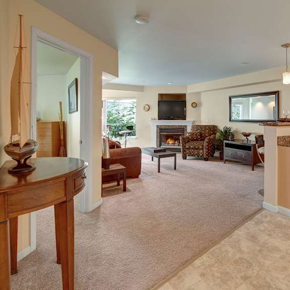 spacious admirals cove living room with beige walls and carpet, featuring a cozy fireplace, brown sofa, patterned armchair, tv, and an adjacent kitchen with a granite countertop