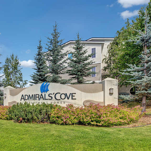 sign for admirals cove apartment homes is surrounded by lush greenery and pine trees. modern buildings are visible in the background under a clear blue sky.