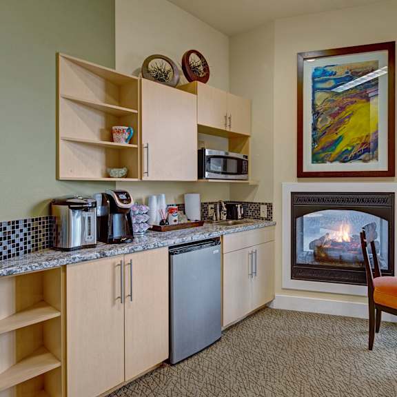 cozy kitchen in the community lounge with light wood cabinets, granite countertop, microwave, and coffee maker. a small fridge, glass table, and lit fireplace add warmth. located at the alpine apartments.