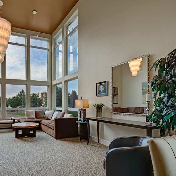 spacious community lounge with large windows, modern chandelier, and neutral tones. features a sectional sofa, round tables, and a decorative mirror. airy and elegant. located at the alpine apartments.