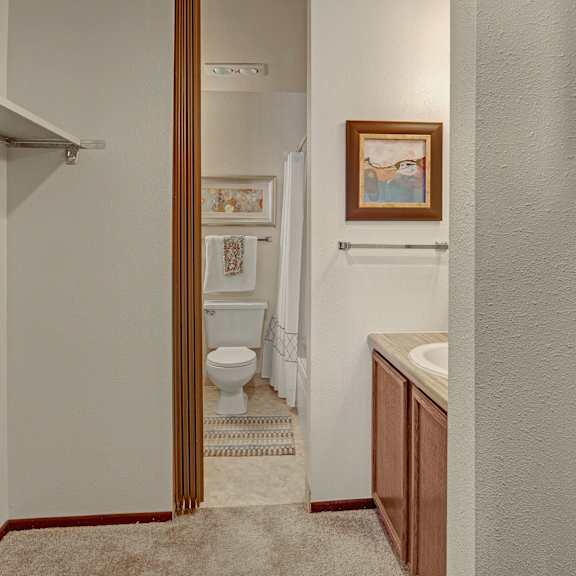 a narrow bathroom with beige walls features a toilet and sink. artwork and a towel hang on the walls, while a striped rug adds subtle color. located at alpine apartments.
