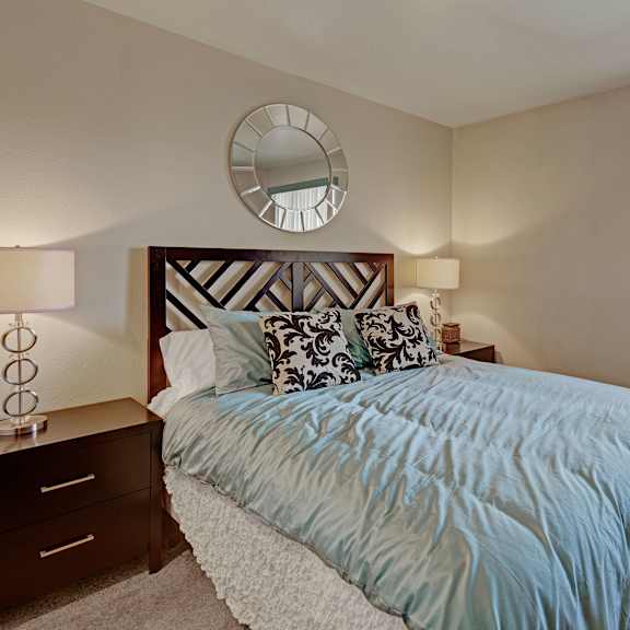 alpine apartments. cozy bedroom with a large bed featuring a blue duvet and patterned pillows. two bedside tables have lamps. a circular mirror hangs on the wall.