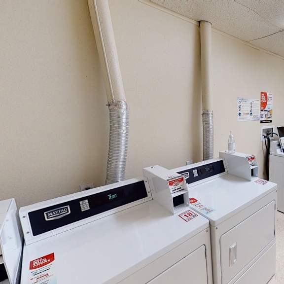 small laundry room with six white washing machines and dryers, beige walls, a door with a window, fluorescent lights, and a red exit sign. amber ridge apartments.