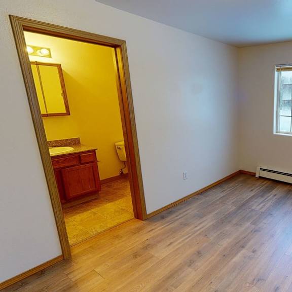 empty bedroom with light wood flooring, cream walls, and a large window showing snowy trees. doorway leads to a brightly lit bathroom. amber ridge apartments.