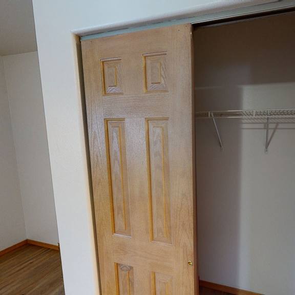 a wooden sliding closet door is partially open, revealing an empty closet with a white wire shelf. the room has light-colored walls and wood flooring. amber ridge apartments.