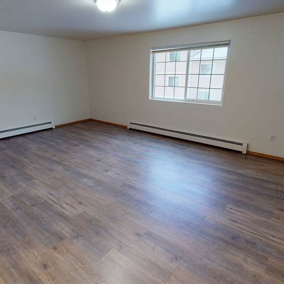empty bedroom with light wood flooring, white walls, and a large window letting in natural light. a ceiling light fixture is visible. cozy and spacious. amber ridge apartments.