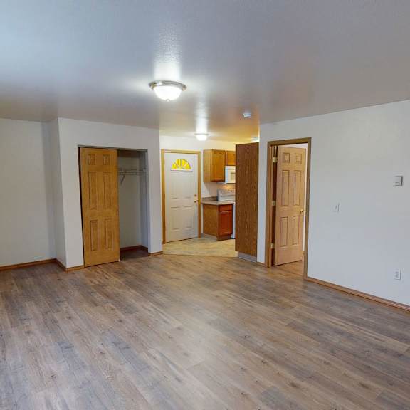 spacious empty livingroom with wooden floors, white walls, and two open wooden doors leading to a kitchen. a closet with sliding doors is visible. calm ambiance. amber ridge apartments.