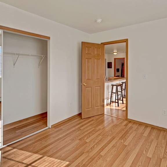 empty bedroom with light wood flooring, white walls, and a mirrored closet with a sliding door. an open wooden door reveals a kitchen with stools. sunlight streams through a window, creating a warm, inviting atmosphere. amber ridge apartments.