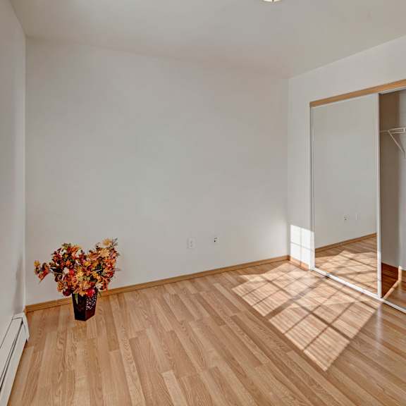 minimalist empty bedroom with hardwood floor and white walls. a large window casts sunlight, highlighting a flower arrangement in a vase. peaceful ambiance. amber ridge apartments.