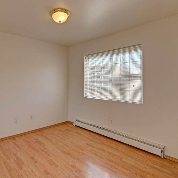 empty bedroom with light wood flooring, cream walls, and a large window with blinds. a ceiling light fixture is centered, creating a serene, clean atmosphere. amber ridge apartments.