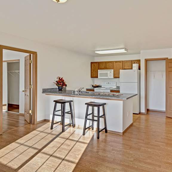 spacious kitchen with light wood cabinets, white appliances, and a breakfast bar with two stools. sunlight streams in, casting shadows on hardwood floor. cozy and inviting. amber ridge apartments.