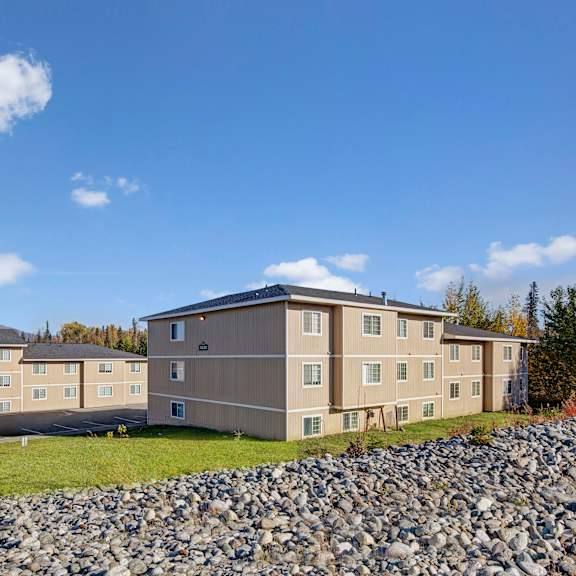 three beige apartment buildings under a clear blue sky, surrounded by trees with autumn foliage and mountains in the distance, convey tranquility. amber ridge apartments.