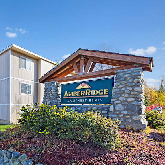 sign for "amber ridge apartment homes" on a stone structure under a clear blue sky. nearby are beige apartment buildings and a parked truck.