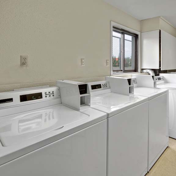 a clean laundromat with four white top-load washing machines lined up. a small window and dark door labeled "boiler room" are visible. bright, orderly atmosphere. arbor pointe apartments.