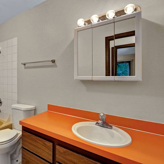 bathroom with yellow bathtub, white tiles, and orange countertop. includes a white toilet, mirror with lights, and wooden cabinets. bright and vintage. arbor pointe apartments.