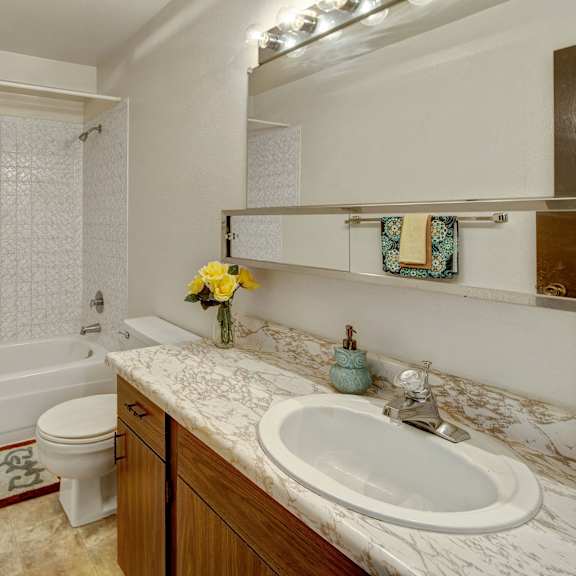 bright bathroom with white tiled bathtub, brown marble sink, and large mirror. yellow roses in vase, towels, and decorative soap add color. cozy and neat. arctic sun apartments.