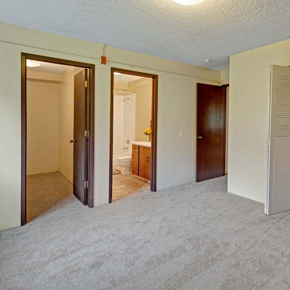 empty bedroom with beige carpet, open closet with white doors on the right, and two wooden doors on the left leading to a bathroom and another room. arctic sun apartments.
