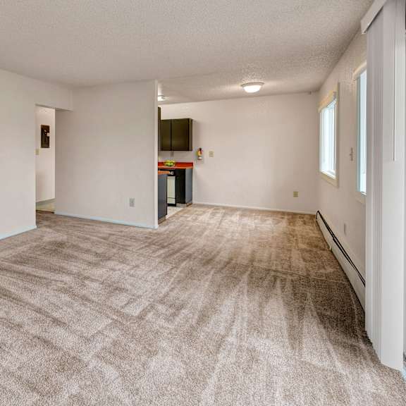 a spacious, neutral-toned living room with beige carpeting, white walls, and an open layout leading to a kitchen. natural light filters through large windows. arctic sun apartments.