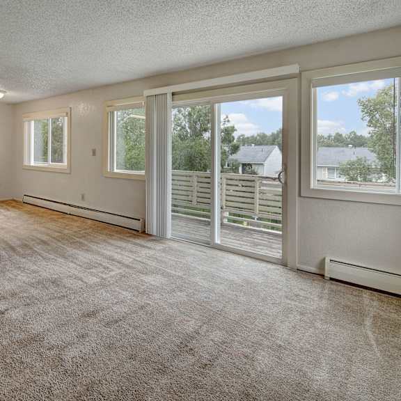 spacious carpeted living room with large windows and sliding glass door leading to a balcony. light gray walls and ceiling create a bright, open feel. arctic sun apartments.