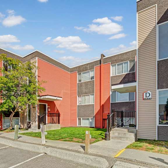 arctic sun apartments building with red and beige siding, two stories, and a "d" sign for units 288-350. trees and parking spaces in the foreground. sky is clear with scattered clouds.