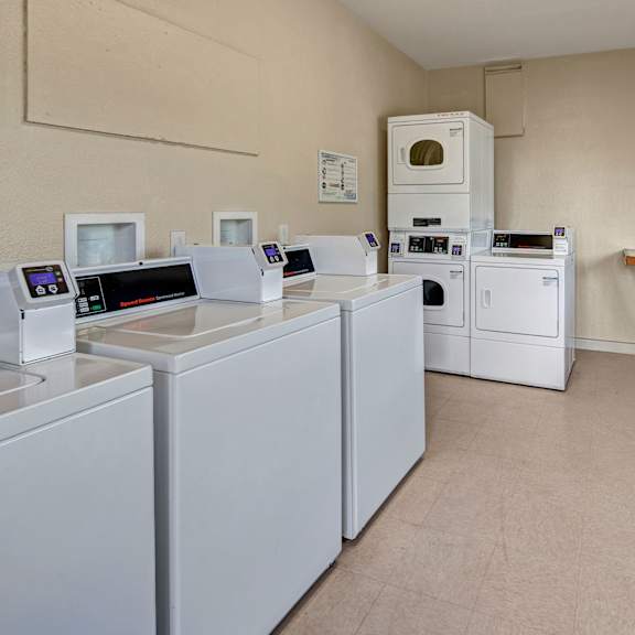 a clean laundry room here at bay arms apartments with three white washing machines and two stacked dryers. a folding table with a vase of flowers adds a welcoming touch.